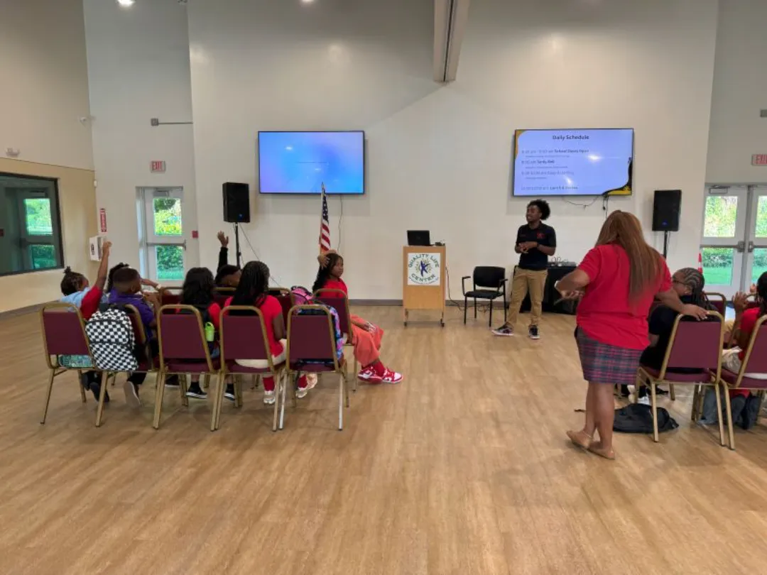Children in colorful clothing raise their hands during a presentation in a spacious room with screens and an American flag